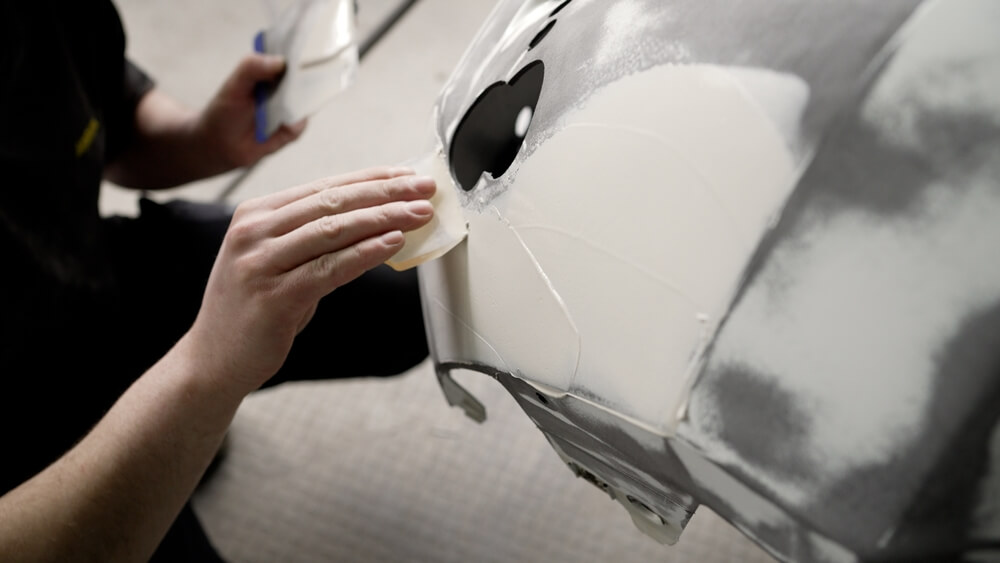 Mechanic sanding bumper damage and applying filler for bumper repair